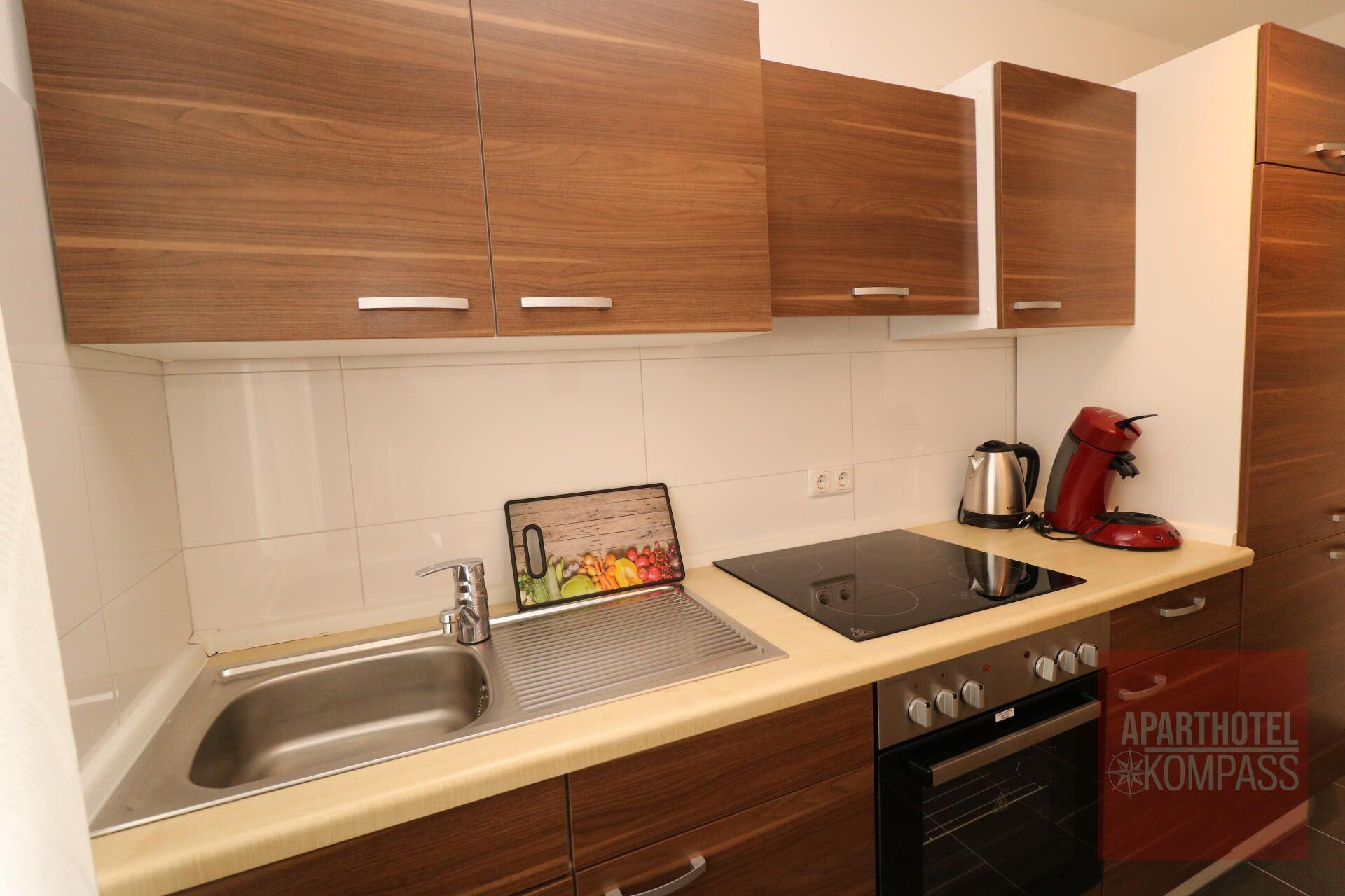 A modern kitchen with wooden cabinets, a stainless steel sink, a built-in stovetop, and a red coffee maker.