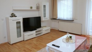 Un salon moderne doté d’un téléviseur à écran plat, de meubles blancs, d’une petite table à manger et d’une fenêtre ornée de rideaux légers.