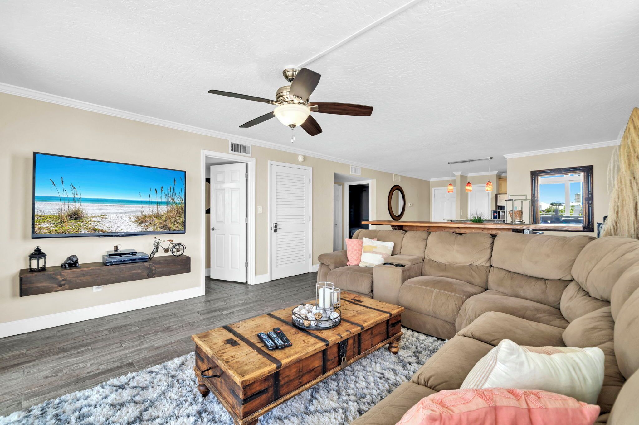 A living room with a large flat-screen TV, a ceiling fan, a wooden coffee table, a beige sofa, and a fluffy rug.