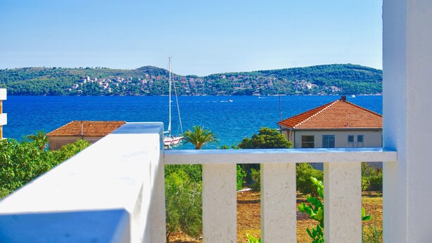 Eine Küstenansicht mit Balkon, der auf das Meer blickt, ein Haus mit rotem Dach und eine in den Hügeln liegende Stadt in der Ferne.