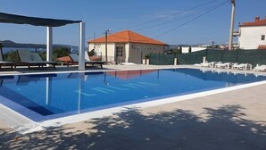 Ein Schwimmbecken mit Liegestühlen und einem überdachten Bereich, umgeben von Gebäuden und blauem Himmel.