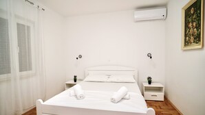 Ein ordentlich bezogenes Bett mit weißen Leinen und zwei gerollten Handtüchern, ein Nachttisch mit einer Pflanze, eine Wand-Klimaanlage und ein gerahmtes Bild an der Wand.