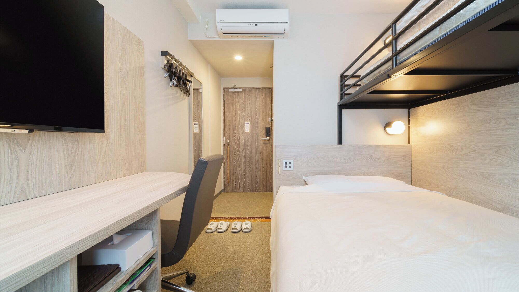 Een compacte hotelkamer met een stapelbed, een bureau met een flatscreen televisie, een stoel en een paar slippers op de vloer.