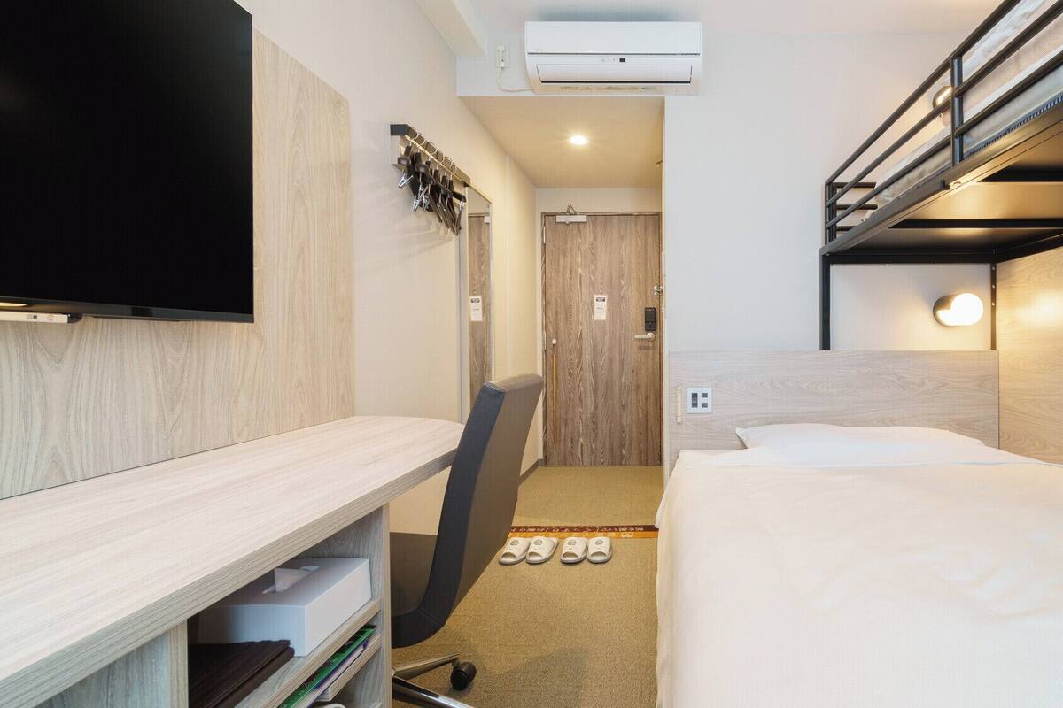 Een compacte hotelkamer met een stapelbed, een bureau met een flatscreen televisie, een stoel en een paar slippers op de vloer.