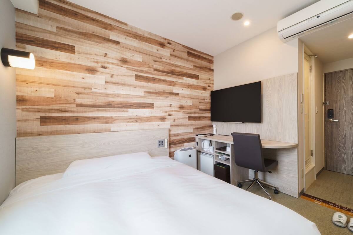 Een moderne hotelkamer met een houten wandpaneel, een bed, een bureau met stoel, een flatscreen televisie en een airconditioner.