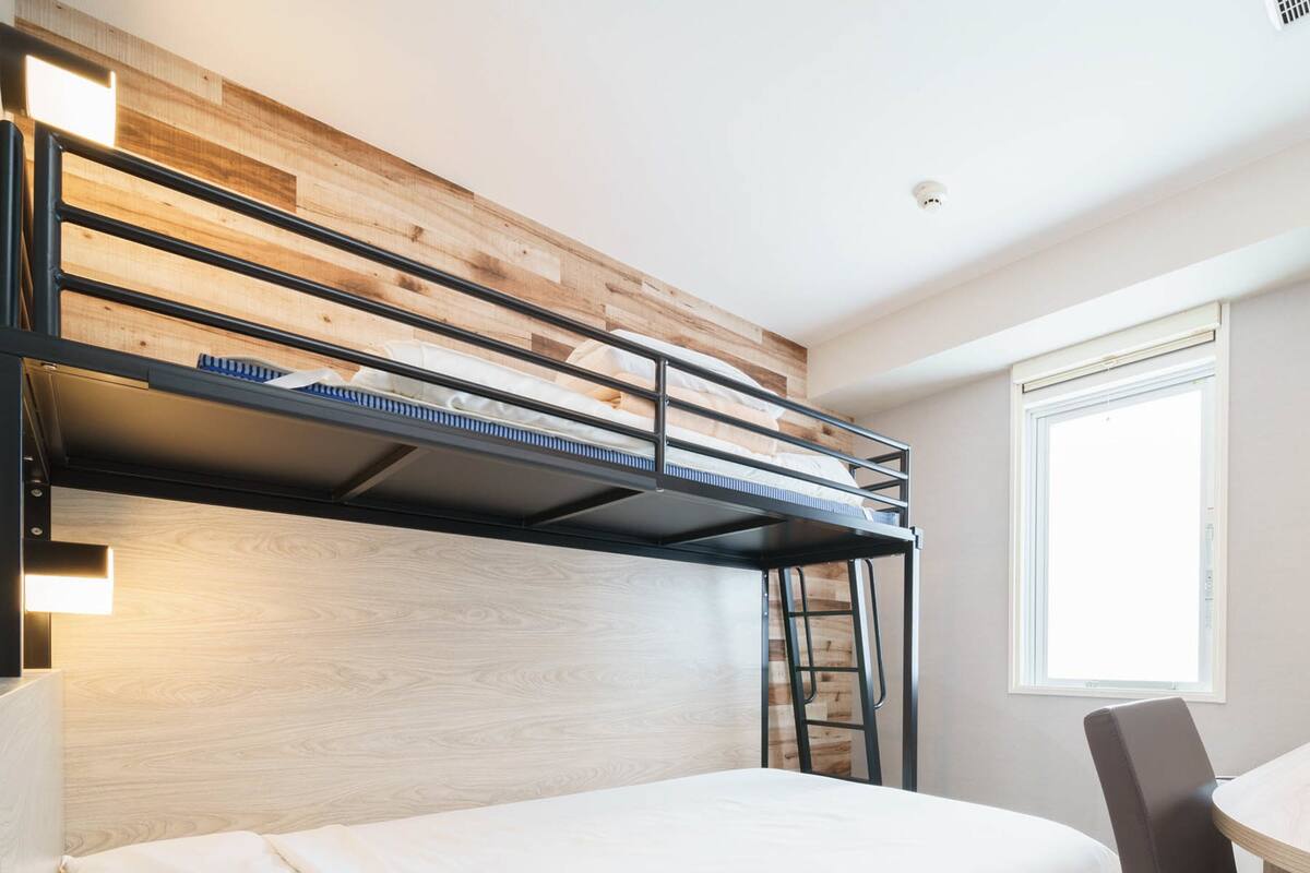 Een stapelbed met een houten hoofdeinde, een bureau met stoel, en een raam met jaloezieën.