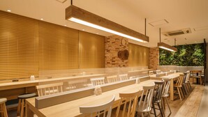 A modern dining area with long wooden tables and chairs, a brick accent wall, and a large window with blinds. - Super Hotel Tokyo JR Kamata Nishiguchi (Tokyo)