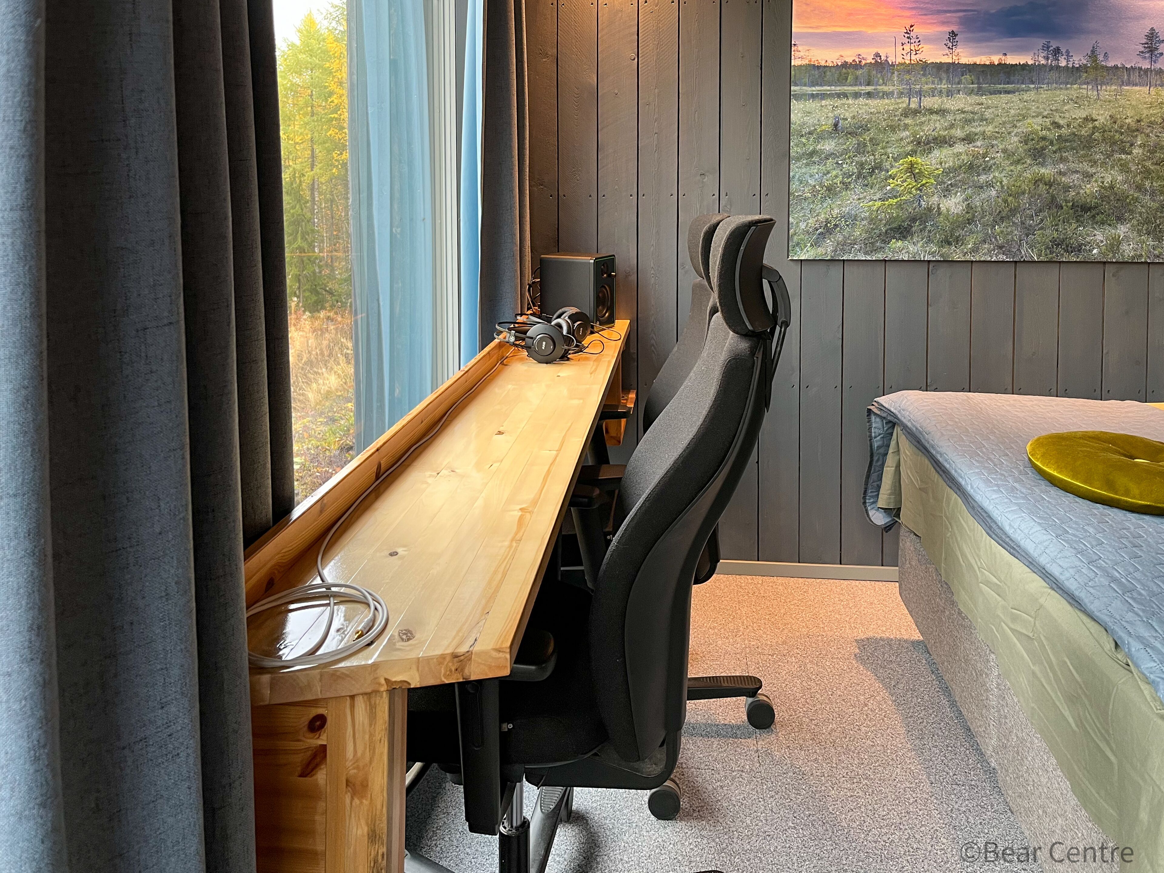 A modern office space with a wooden desk, black office chair, and a view of a field through a window.