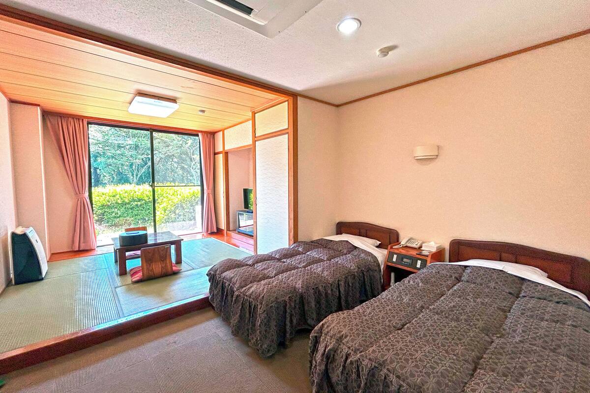 Une chambre d’hôtel avec deux lits, une petite table et une vue sur la verdure, accessible par une porte coulissante.