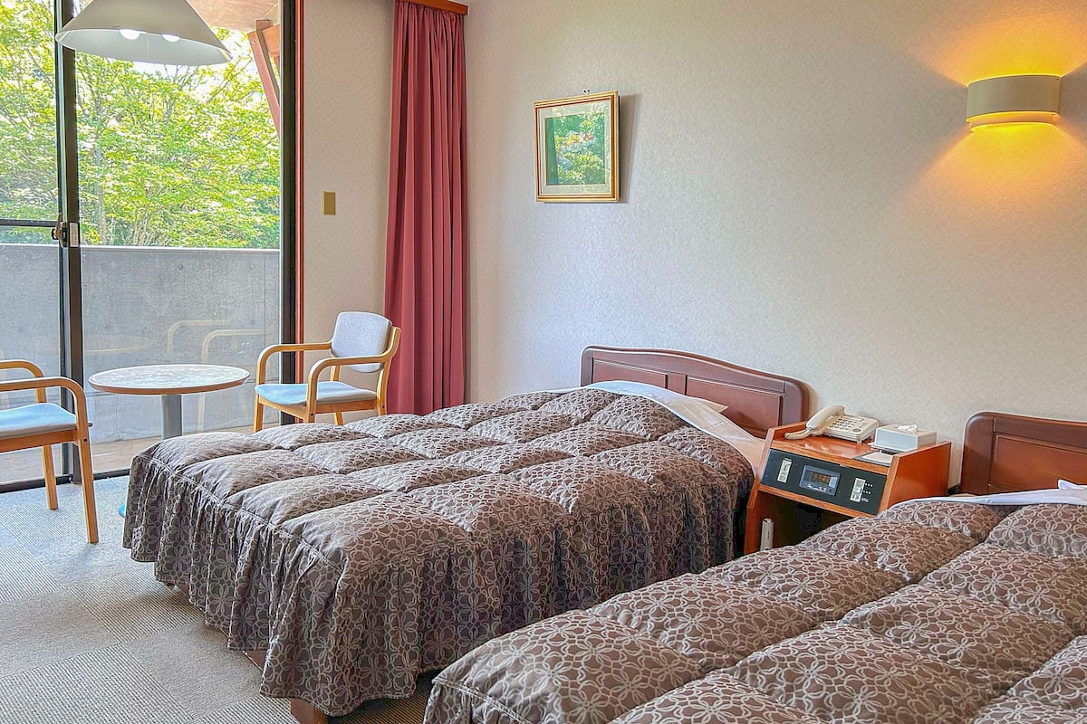 Une chambre d’hôtel avec deux lits, une petite table avec un téléphone, une chaise et une fenêtre donnant sur des arbres.