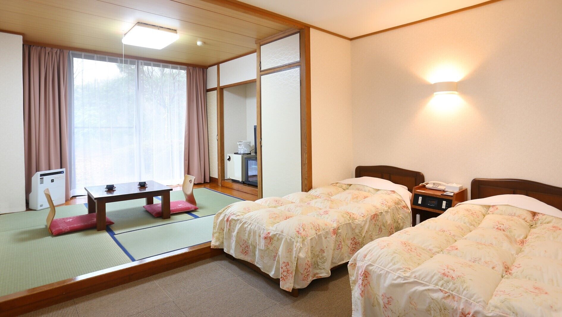 Une chambre d’hôtel avec un sol recouvert de tatamis, un lit de style japonais traditionnel, une petite table avec une chaise et un paravent coulissant de type shoji.