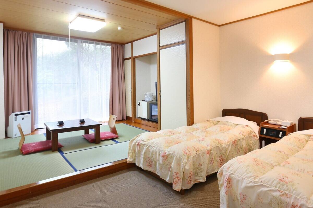 Une chambre d’hôtel avec un sol recouvert de tatamis, un lit de style japonais traditionnel, une petite table avec une chaise et un paravent coulissant de type shoji.