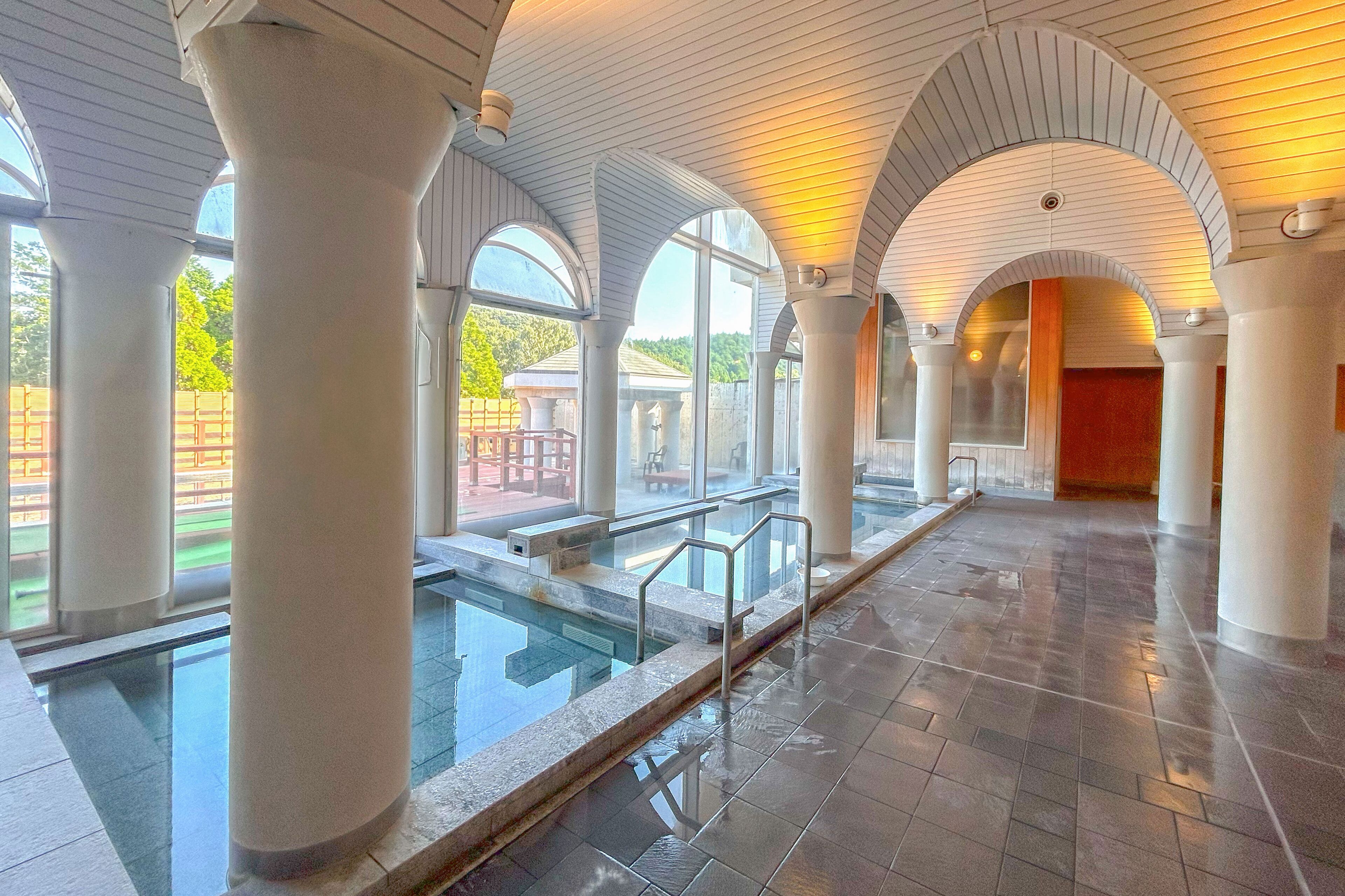 A spacious indoor swimming pool area with clear glass railings, large white columns, and arched ceilings.