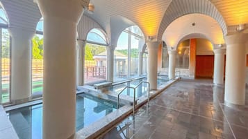 A spacious indoor swimming pool area with clear glass railings, large white columns, and arched ceilings.