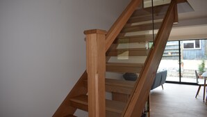 Uma escada de madeira com guarda-corpos de vidro leva a uma área de estar com um sofá e uma mesa de jantar.