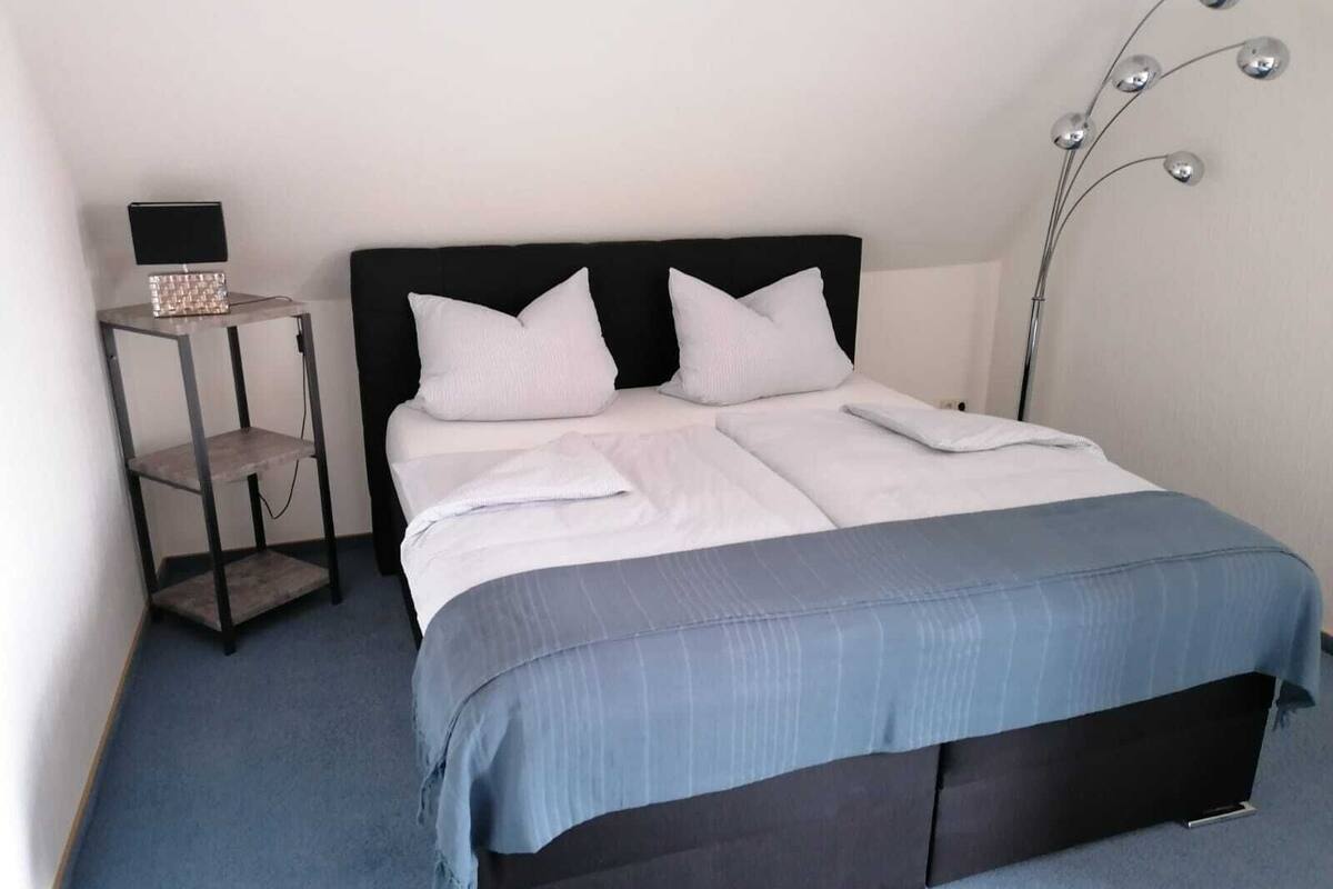 A neatly made bed with white linens and a blue blanket, a black headboard, and a bedside table with a lamp.