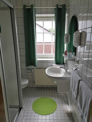 A bathroom with a toilet, sink, and bathtub, a green mat on the floor, and a window with curtains. - Hotel Biedendieck (Warendorf)