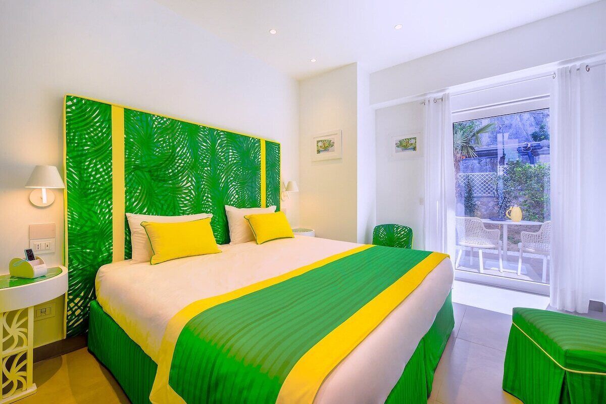 Een hotelkamer met een bed, een nachtkastje, een groen decoratief hoofdeinde en uitzicht buiten door een groot raam.