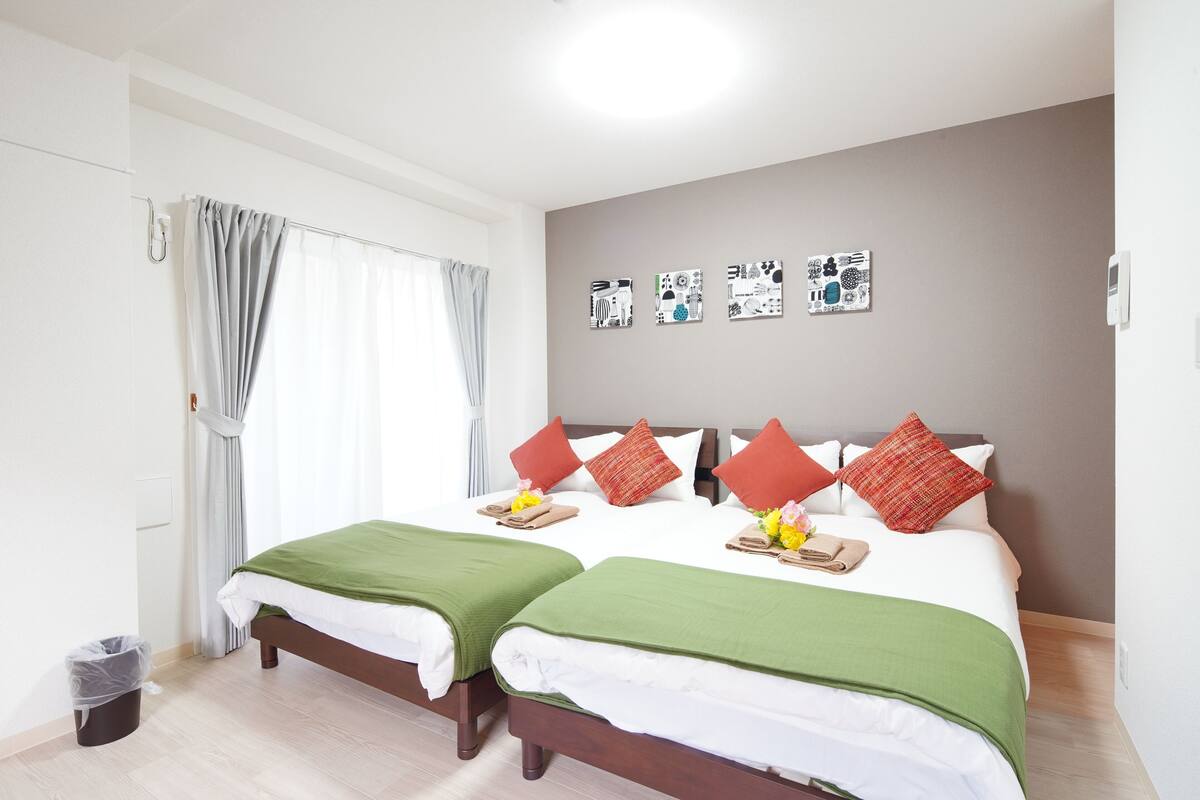 Une chambre d’hôtel avec deux lits, un mur gris orné d’œuvres d’art, une fenêtre avec des rideaux et une poubelle.