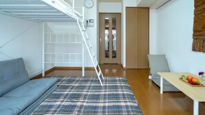 A compact living space with a bunk bed, a sofa, a plaid blanket, a wooden table with fruit, and a door leading to another room. - Liv City Shinjuku Ⅱ 504 (Tokyo)