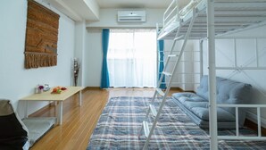 A room with a bunk bed, a desk, a chair, a table with fruits, a wall hanging, and a window with curtains. - Liv City Shinjuku Ⅱ 504 (Tokyo)