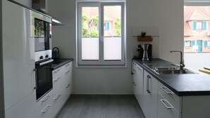Eine moderne Küche mit weißen Schränken, einer schwarzen Arbeitsplatte, einem eingebauten Ofen, einem Fenster mit Jalousien und Blick auf eine Wohngegend.