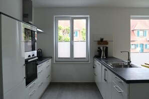 Eine moderne Küche mit weißen Schränken, einer schwarzen Arbeitsplatte, einem eingebauten Ofen, einem Fenster mit Jalousien und Blick auf eine Wohngegend.