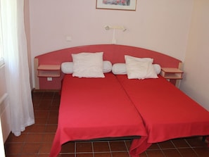 A twin bed with a red bedspread and white pillows, a bedside table, and a small shelf. - Small Studio No. 5 with Ocean View (Bormes-les-Mimosas)