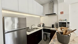 Una cocina moderna con un refrigerador de acero inoxidable, un microondas y un tostador. Hay un lavamanos con grifo, un cubo de basura y una cesta de madera para cucharas sobre la encimera.
