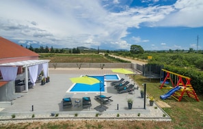 Un área de piscina en la azotea con sillones de descanso, sombrillas y un parque infantil.