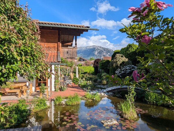 Une maison en bois avec un jardin, un étang parsemé de nénuphars et une montagne en arrière-plan.