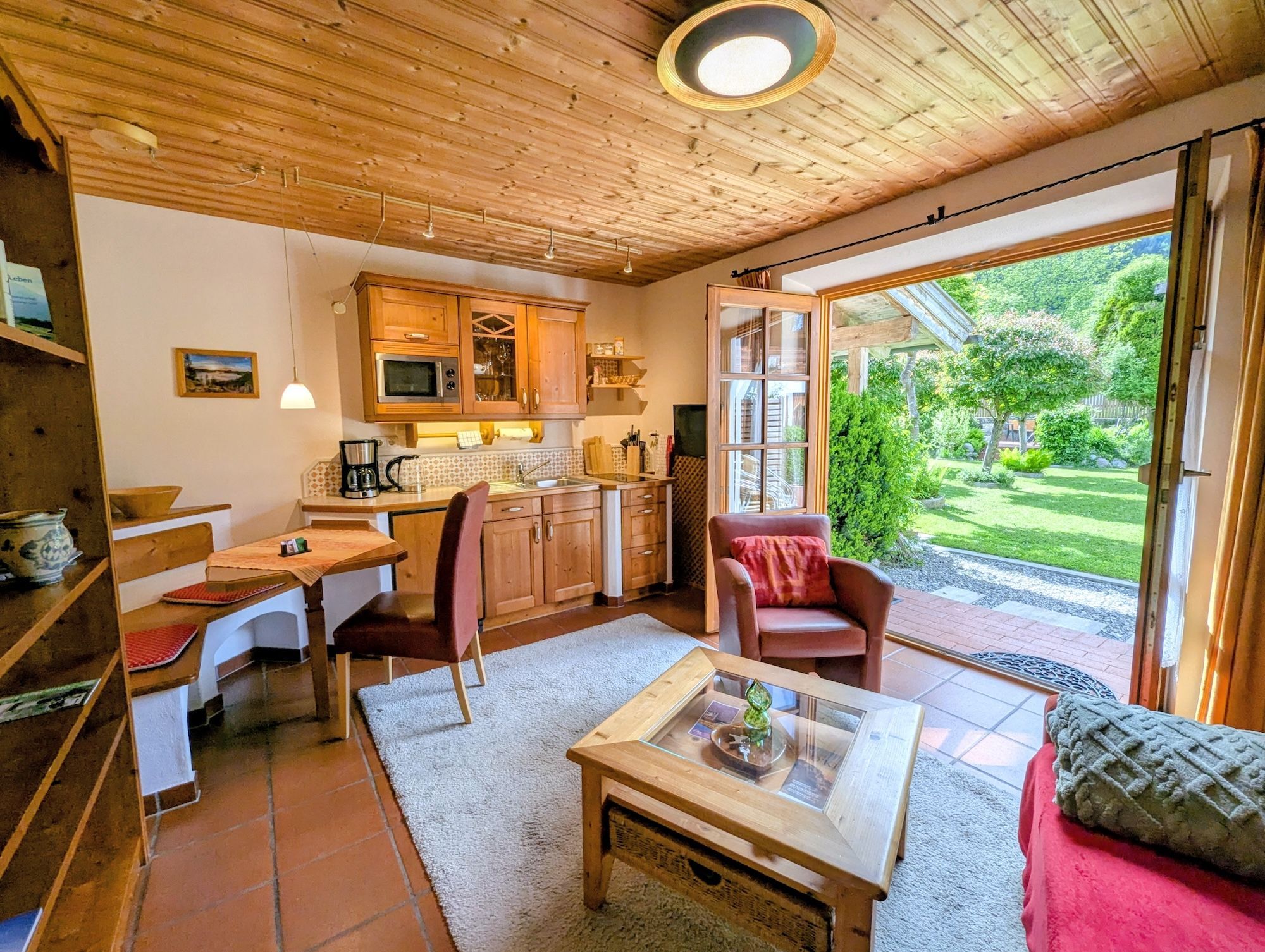 A cozy interior with a wooden ceiling, a kitchen with a microwave, a dining area with a table and chairs, and a view of a garden through a glass door.