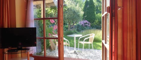 Habitación con televisor sobre una mesa de vidrio, puerta corrediza de vidrio que da a un jardín con sillas y una mesa afuera.