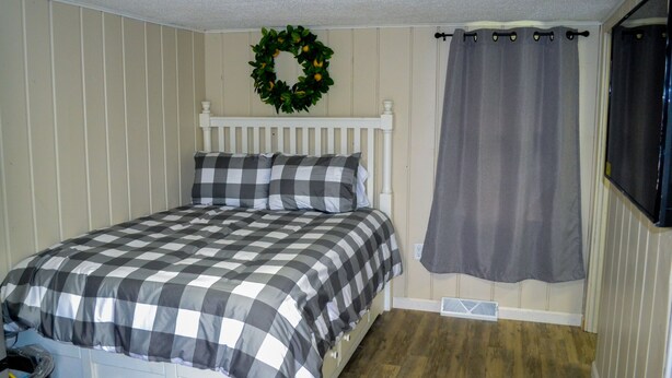 Ein Schlafzimmer mit kariertem Bettbezug, einem weißen Kopfteil, einem grauen Vorhang und einem grünen Kranz an der Wand.