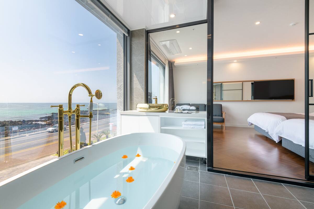 A modern bathroom with a large bathtub, a shower, and a vanity. It has a view of the ocean and a road.