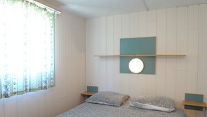 Un dormitorio con una cama, dos mesitas de noche, una lámpara de pared y un ventanal con cortinas.