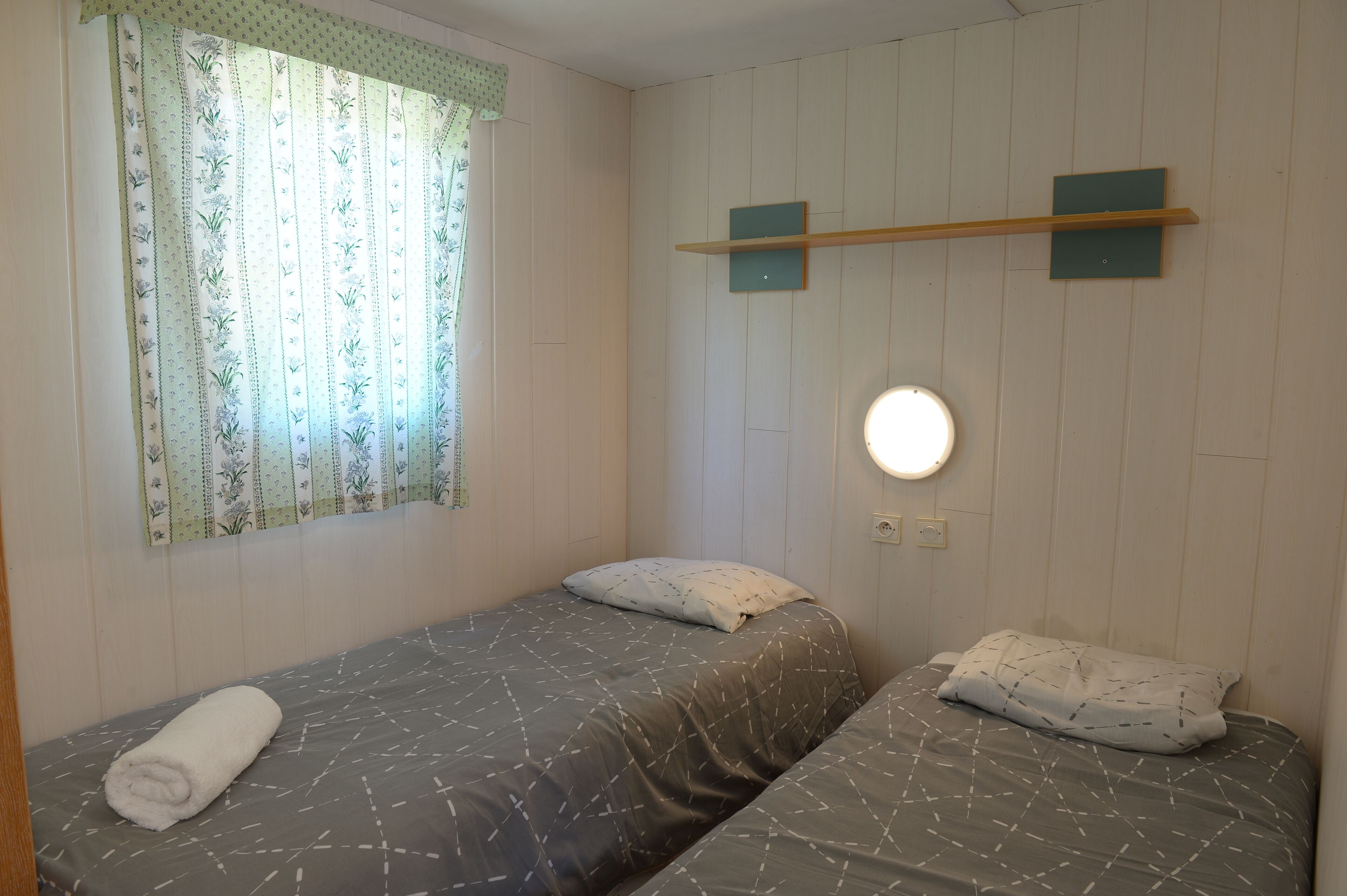 Una habitación pequeña y limpia con dos camas individuales, una ventana con cortinas florales y una estantería en la pared.