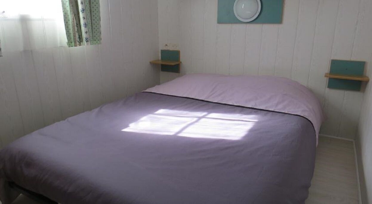 A single bed with a purple bedspread in a room with white walls and a window.