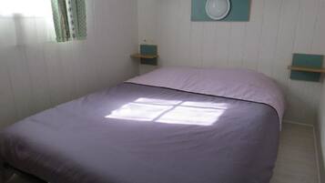 A single bed with a purple bedspread in a room with white walls and a window.