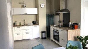 A compact kitchen with white cabinets, a sink, a stove, and a microwave. There is a trash bin and a clock on the wall. - Etoile de Mer : T2 Avec Jardin, Accessible aux Personnes à Mobilité Réduite, (Sète)