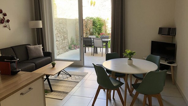 A modern living room with a sofa, coffee table, dining table, chairs, and a TV. There is a sliding glass door leading to an outdoor area with a table and chairs. - Etoile de Mer : T2 Avec Jardin, Accessible aux Personnes à Mobilité Réduite, (Sète)