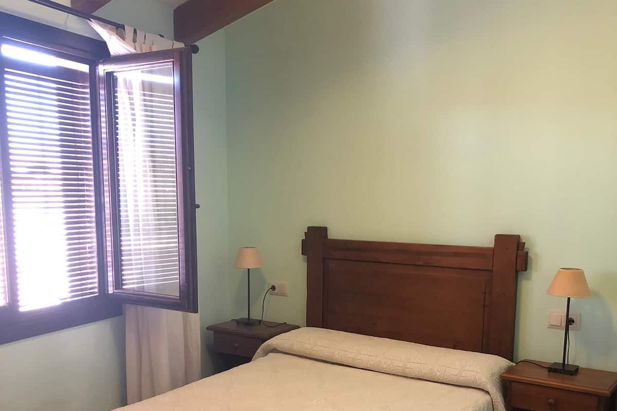 A bedroom with a bed, two bedside tables, a window with blinds, and a lamp on each table.