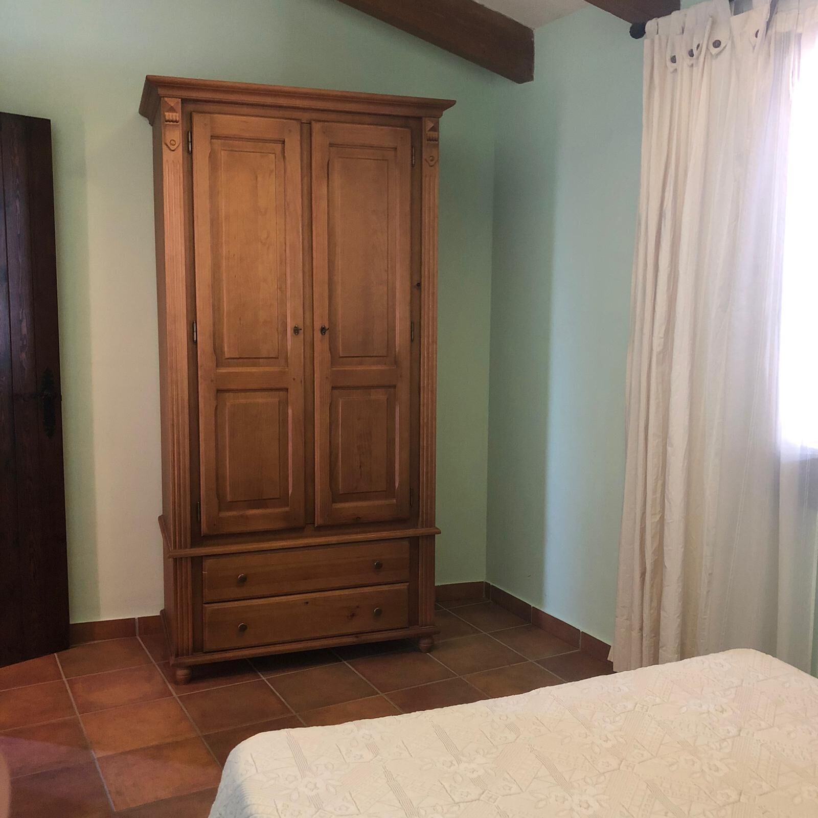 Un dormitorio con un armario de madera, una cama con cubrecama estampado y una ventana con cortinas.