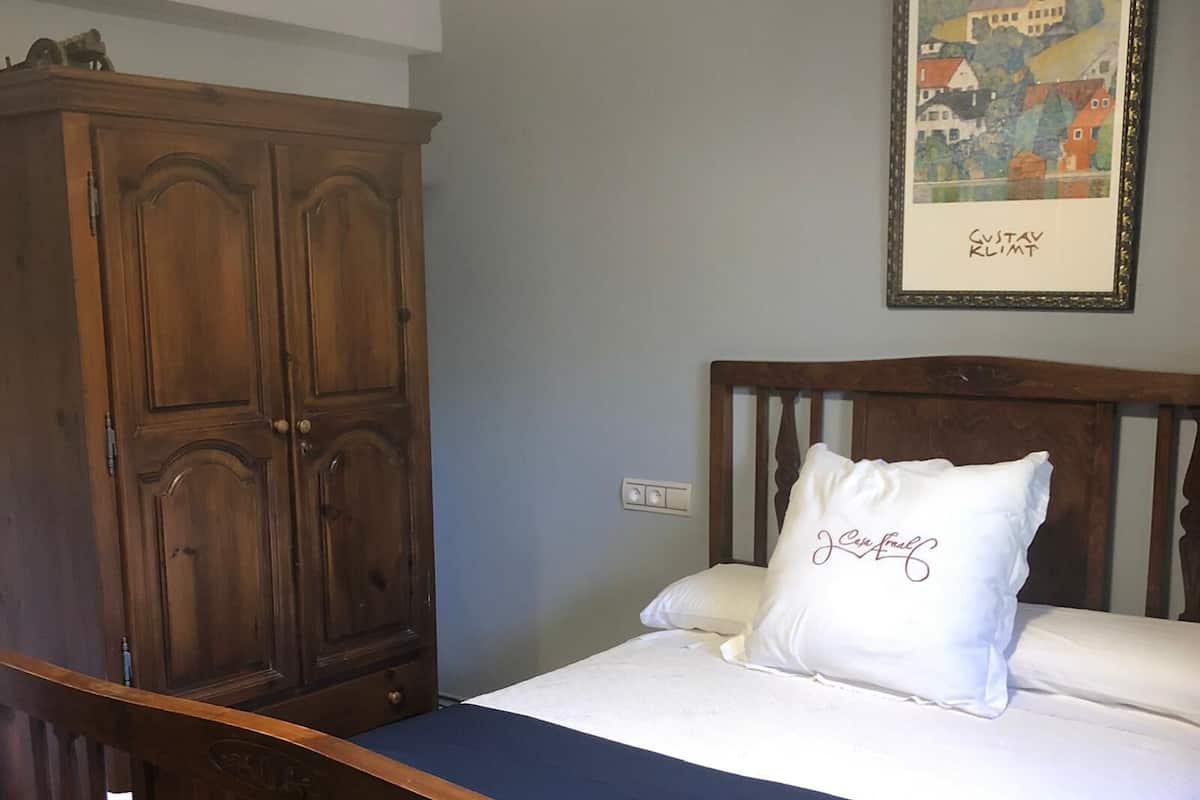 A bedroom with a wooden bed frame, a white mattress, and a blue bed skirt. There is a wooden wardrobe with a mirror and a framed picture on the wall.