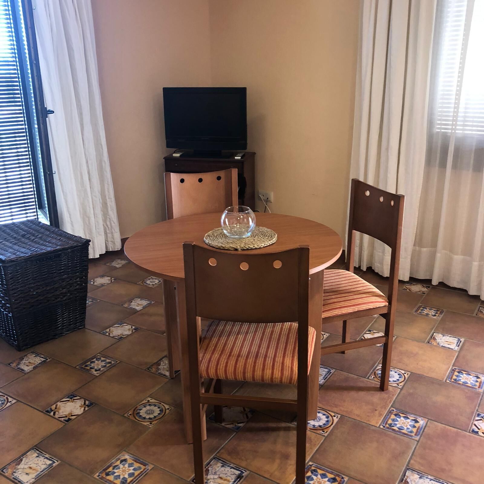 A dining area with a round table, two chairs, a woven placemat, a television on a stand, and patterned floor tiles.
