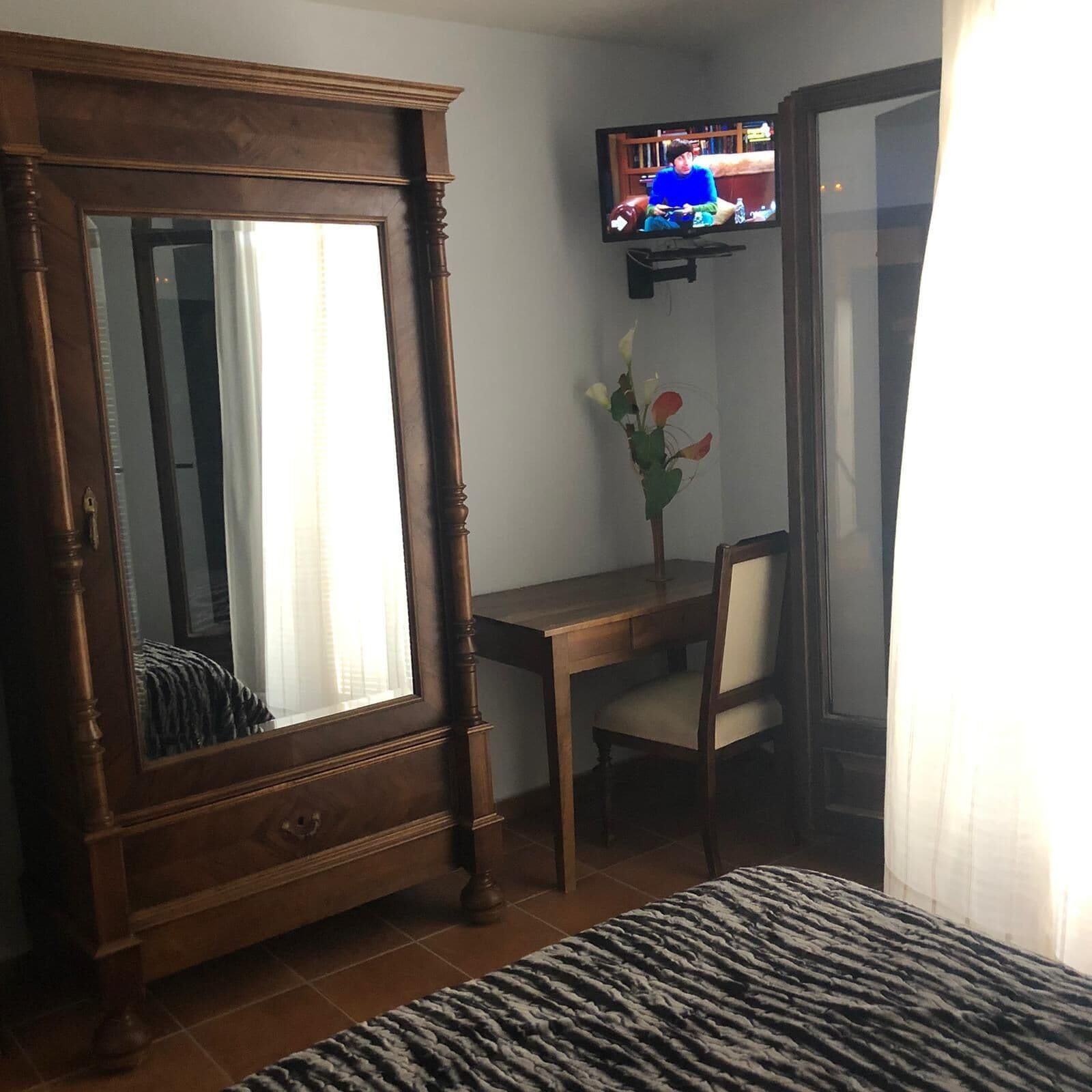 Ett sovrum med en garderob i trä, ett skrivbord med en stol, en tv som är monterad på väggen och en säng med ett mönstrat överkast.