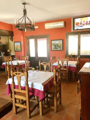 Un comedor con mesas y sillas de madera, una araña y un televisor en la pared.