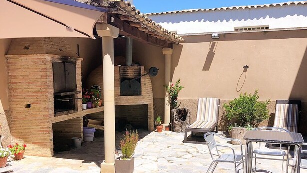 Un patio exterior con suelo de piedra, un horno de ladrillo, sillas de metal y una mesa.