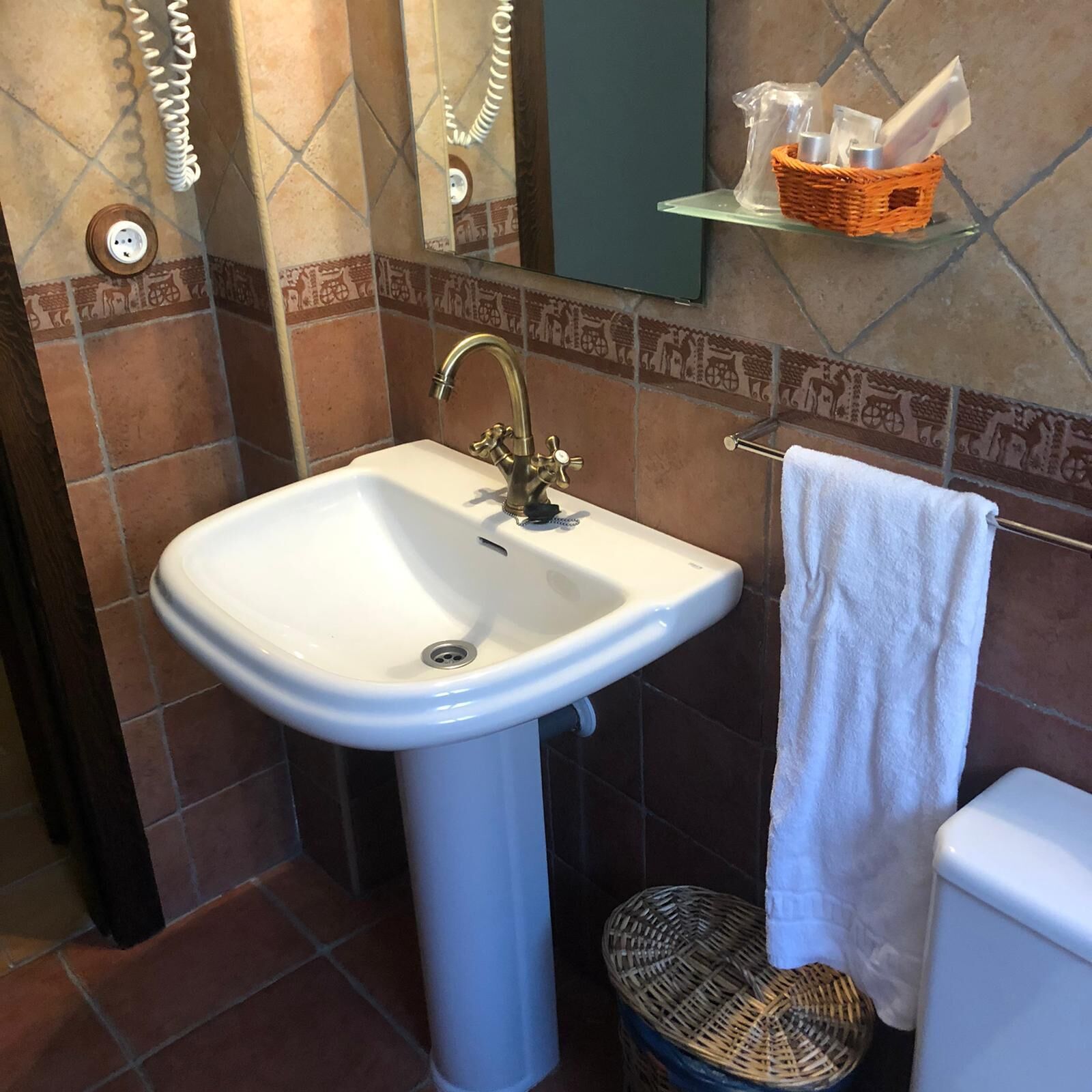 Un baño con un lavamanos blanco de pedestal, un perchero con una toalla blanca, una cesta de mimbre en un estante y un inodoro.