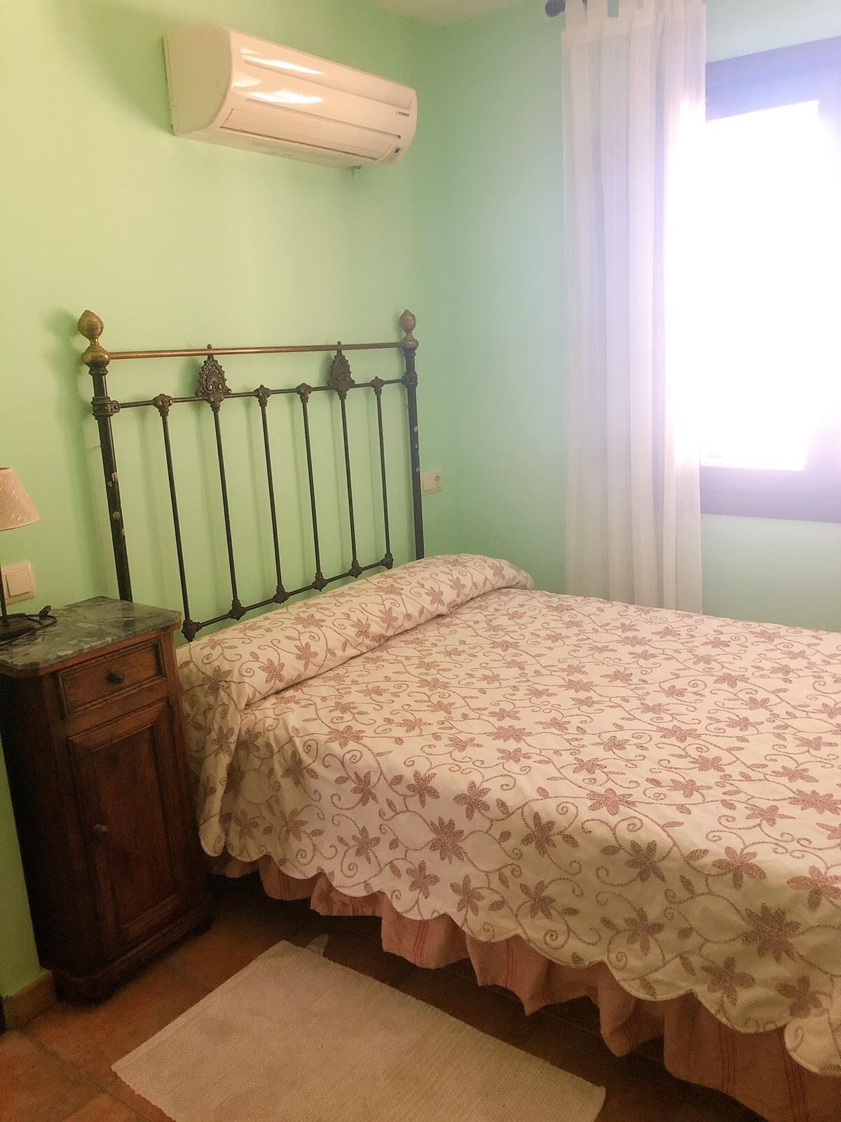 Une chambre à coucher comprenant un lit, une table de chevet, une commode, un climatiseur et une fenêtre avec des rideaux.